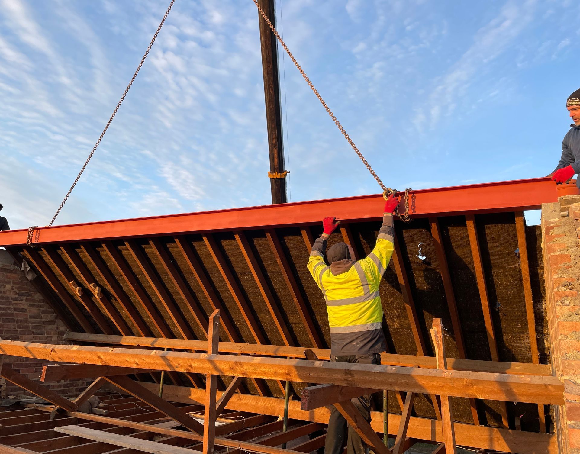 Workers installing a red steel beam on a building. They're wearing safety gear, and it's a sunny day.