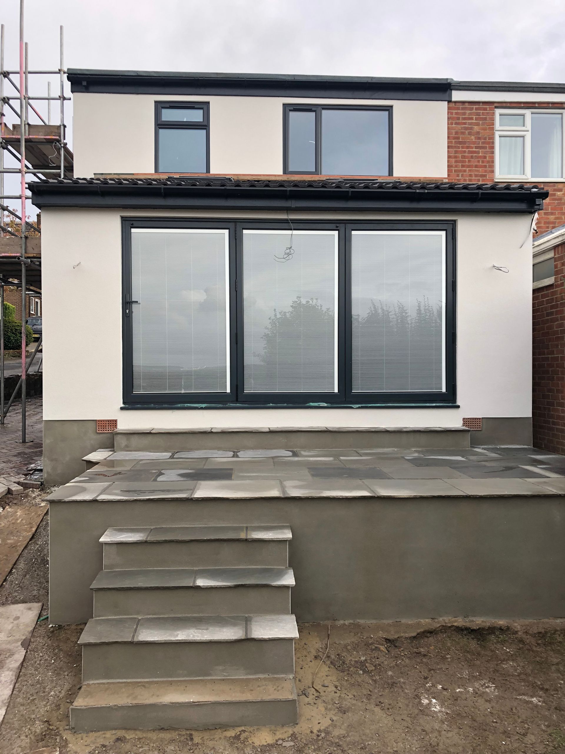 Rear view of a house extension with gray framed windows and a patio with steps. Gray walls and scaffolding.
