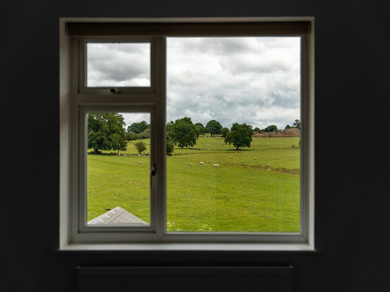 Window framing a green field with trees and a cloudy sky.