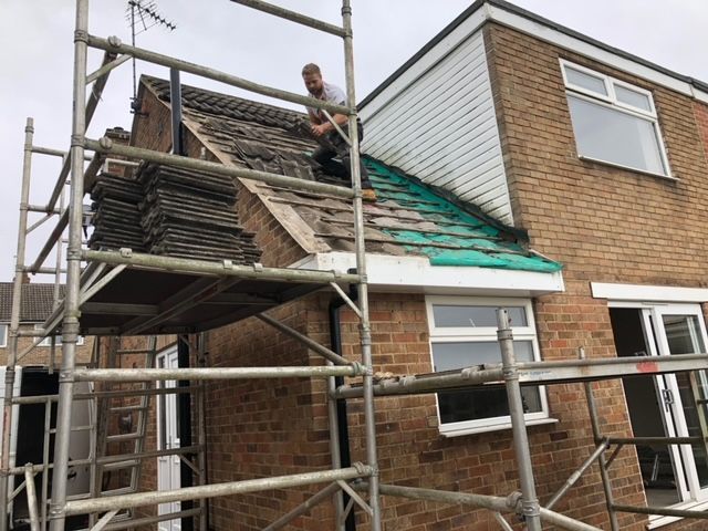 Man on scaffolding, removing roof tiles from a brick house with white trim.