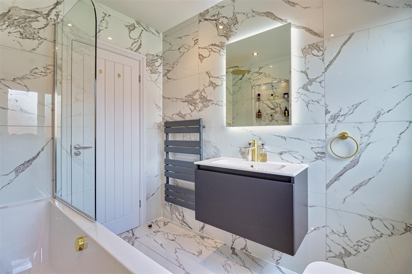Modern bathroom with marble-effect tiles, gray vanity, illuminated mirror, and gold fixtures.
