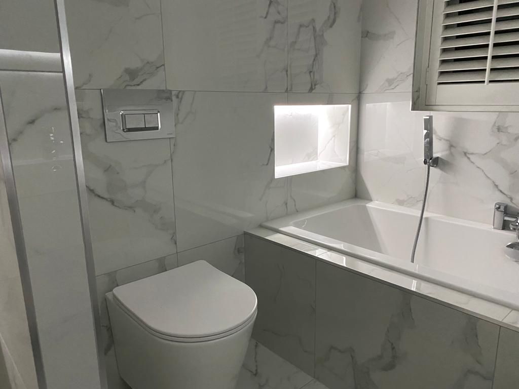 Bathroom with marble-effect tiles, toilet, bathtub with niche, and shuttered window.
