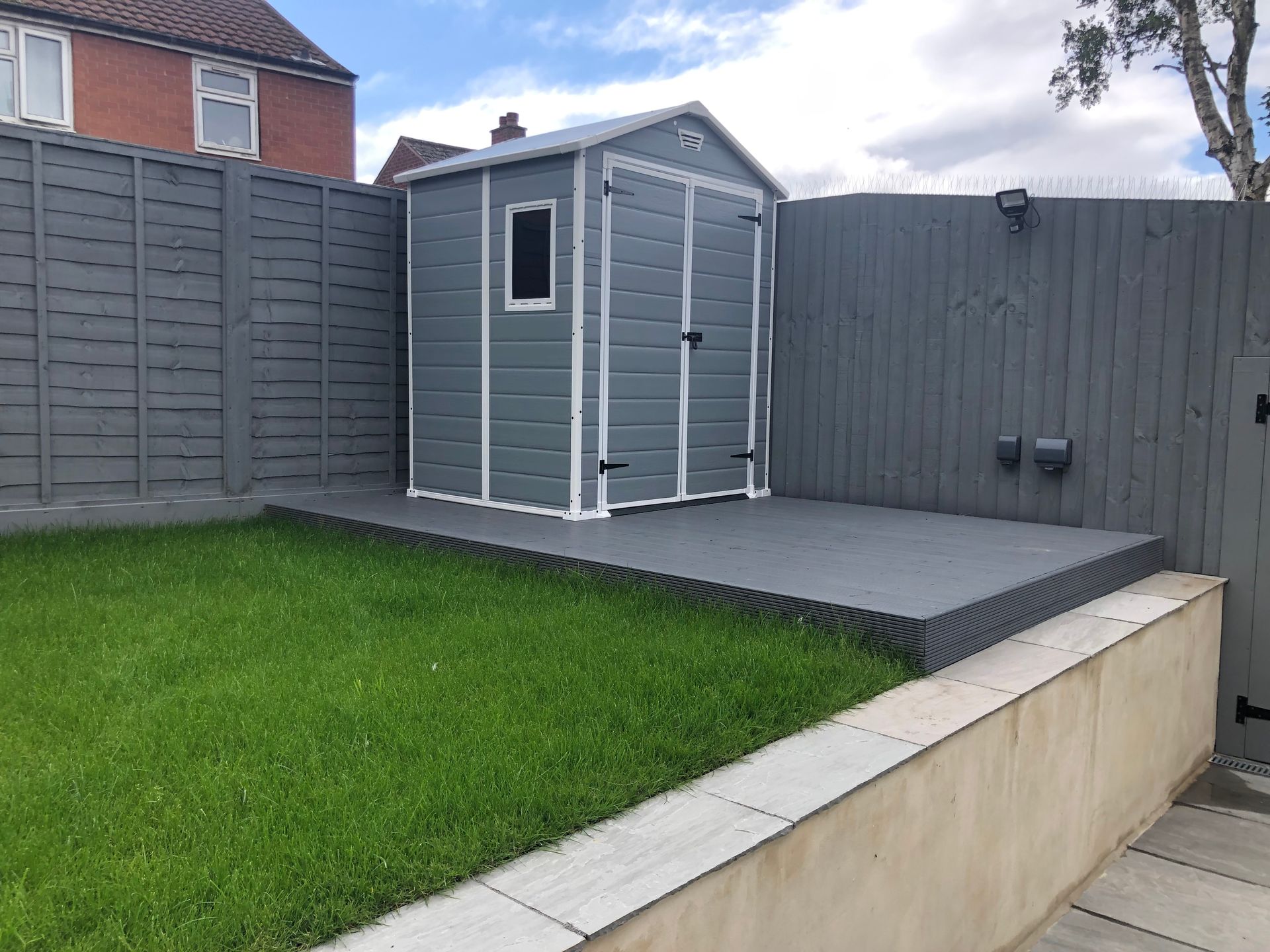 Gray shed on a gray platform, bordered by a gray fence and green lawn.