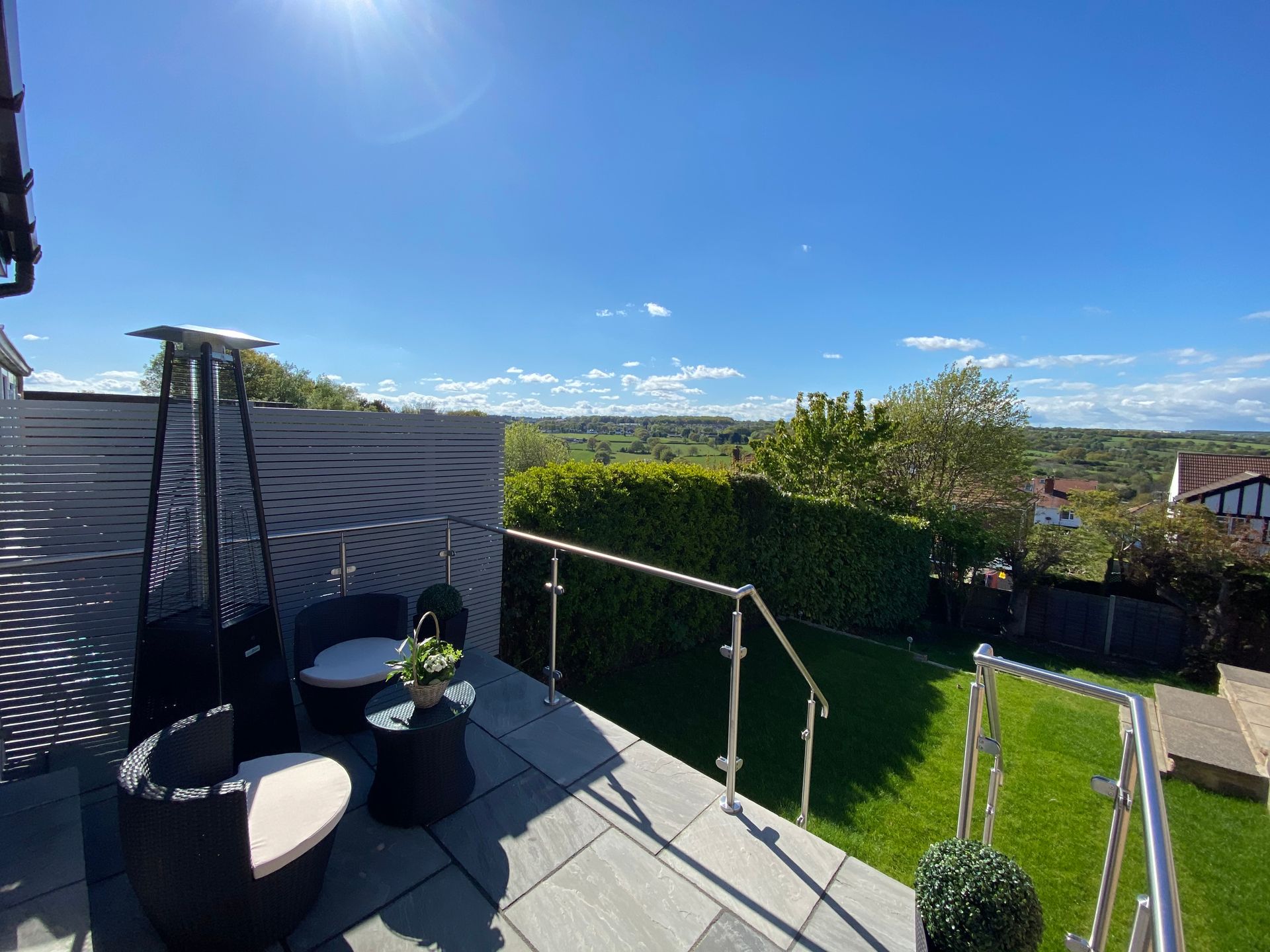 Patio with black wicker chairs, table, heater, green grass, and a scenic view under a blue sky.
