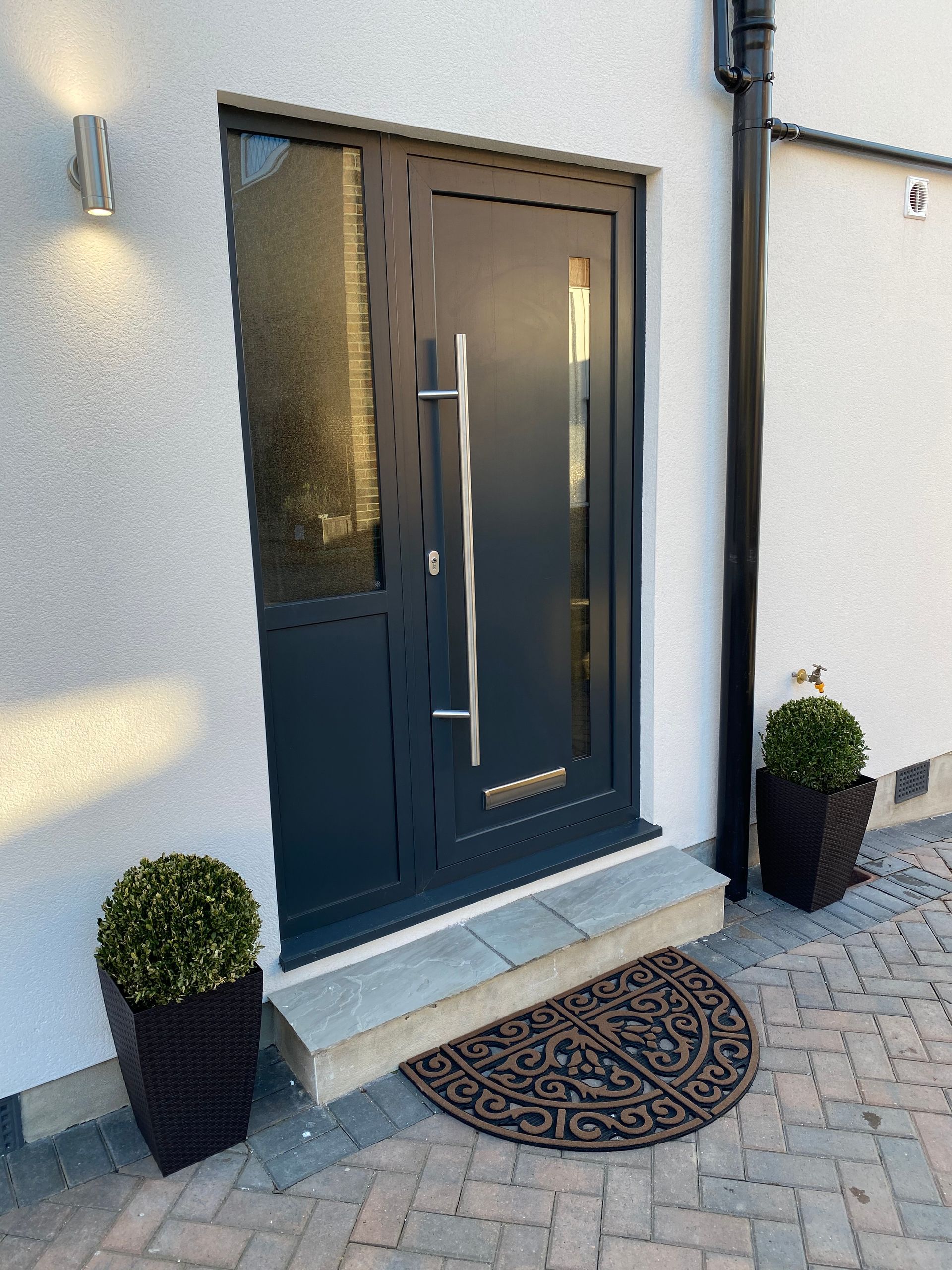 Dark gray front door with sidelight, stainless steel handle. Two planters with topiary on brick pavers.