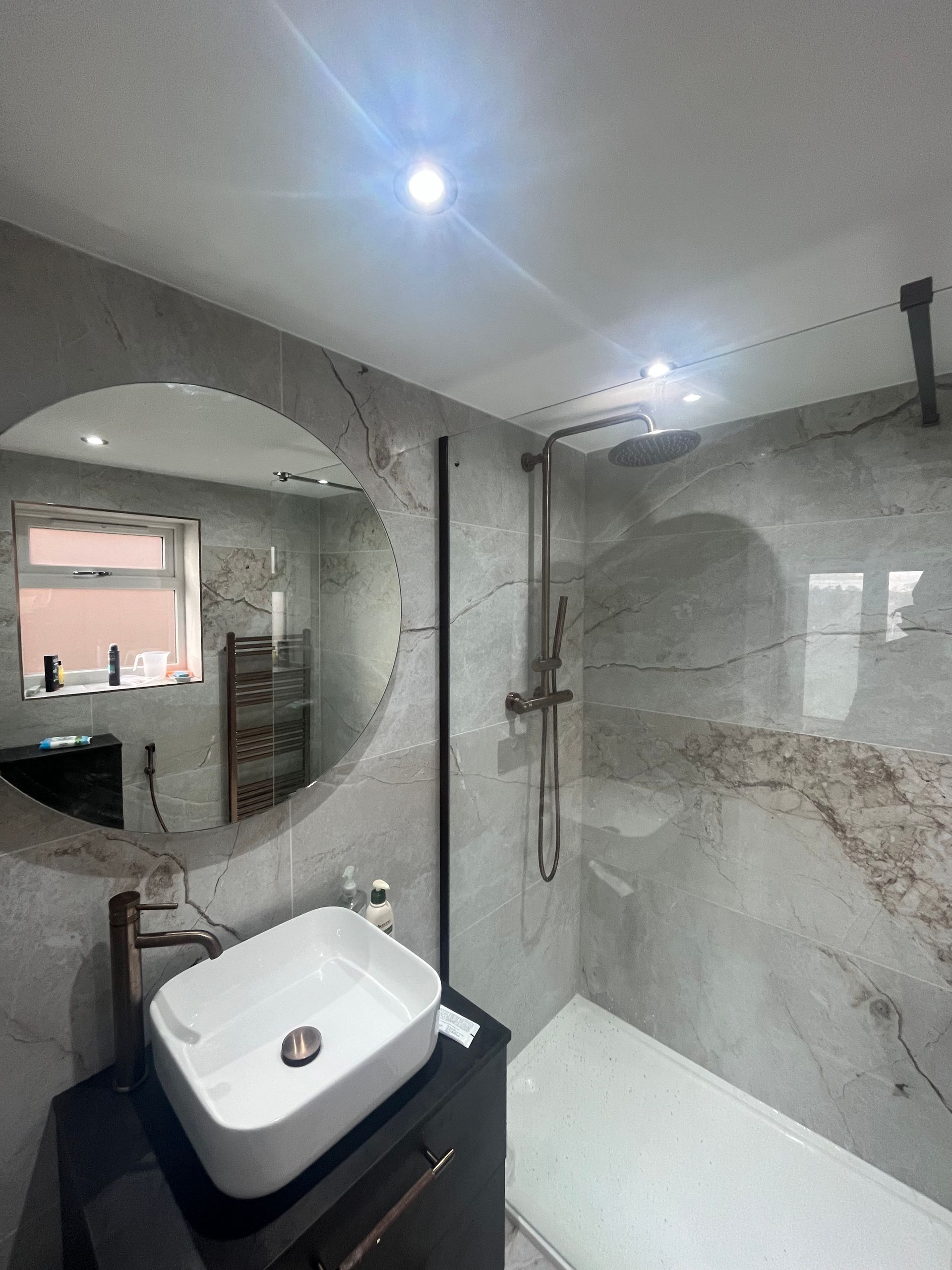 Modern bathroom with a marble-look tiled shower, round mirror, and a floating sink.