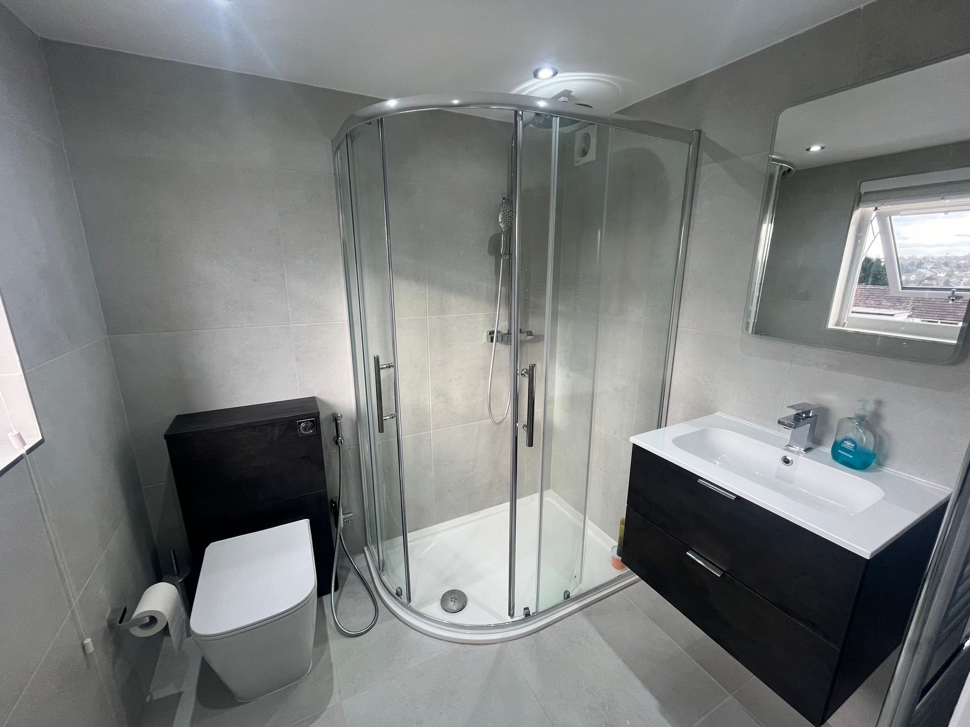 Modern bathroom with a shower, toilet, vanity, and mirror. Grey walls, black vanity, and white fixtures.