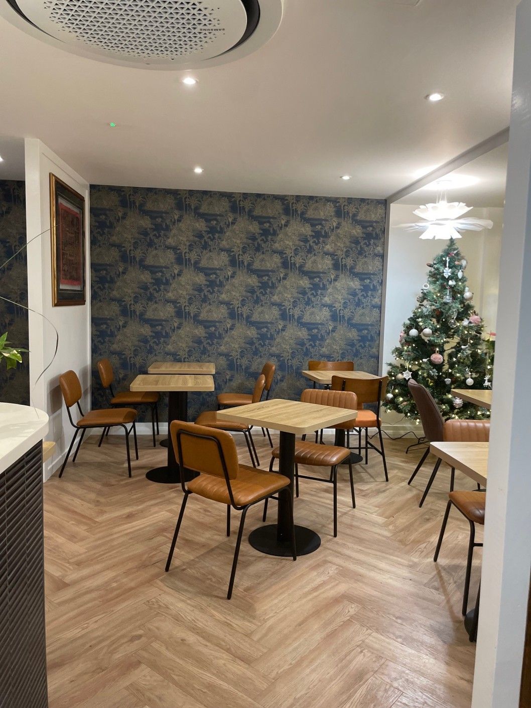Interior of a cafe: tables and chairs, blue floral wallpaper, Christmas tree, wooden floors, and ceiling fan.
