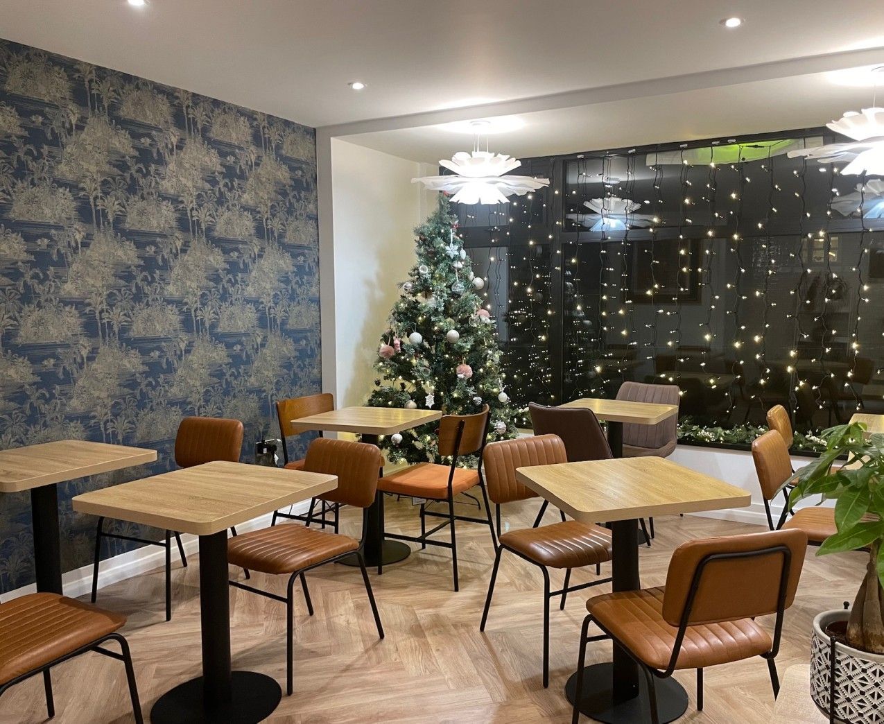 Cafe interior with tables, chairs, blue floral wallpaper, Christmas tree, and string lights.