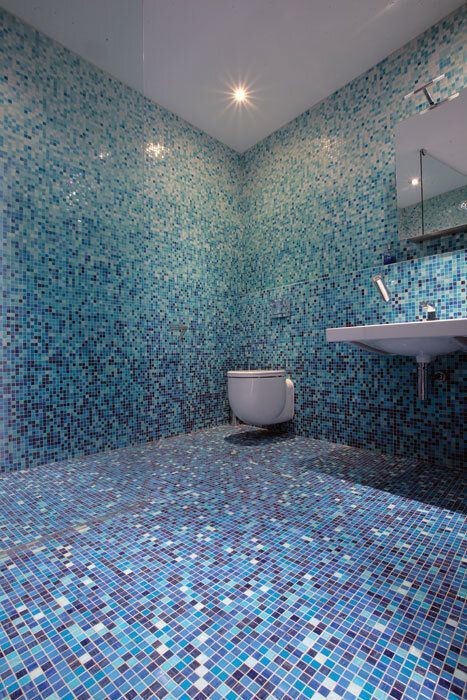 Blue mosaic-tiled bathroom, toilet and sink, walls and floor, bright overhead light.
