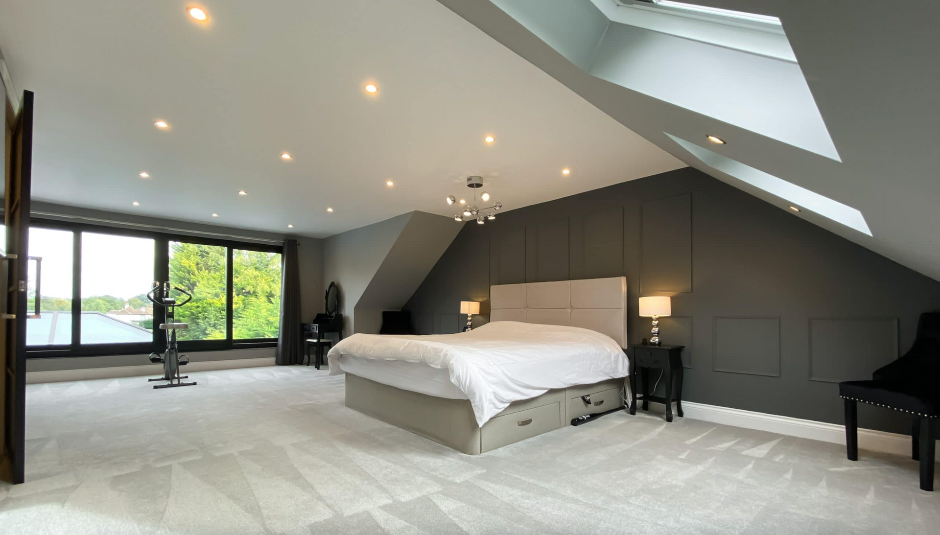 Spacious modern bedroom with large windows, gray walls, and a bed, with natural light from a skylight.