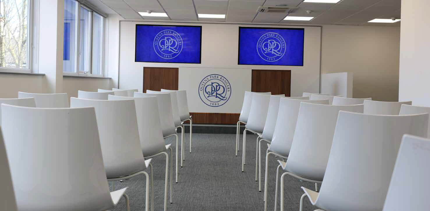 Empty conference room with rows of white chairs facing a podium and blue presentation screens