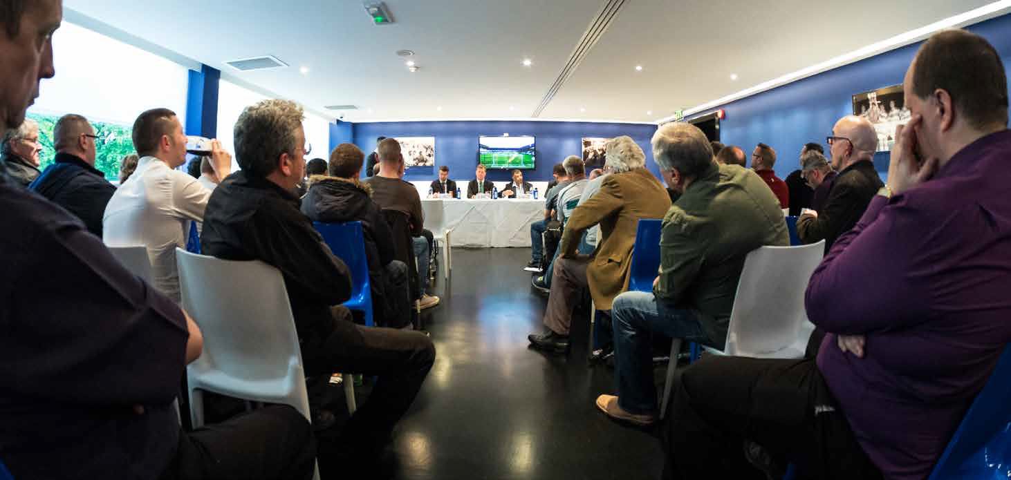 Audience seated in a conference room, watching a presentation on large screens at the front.