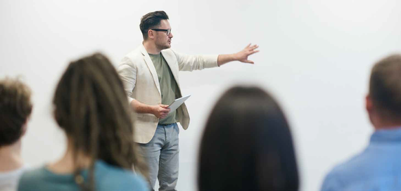 Presenter gesturing at a screen in a meeting room, with audience members blurred in the foreground