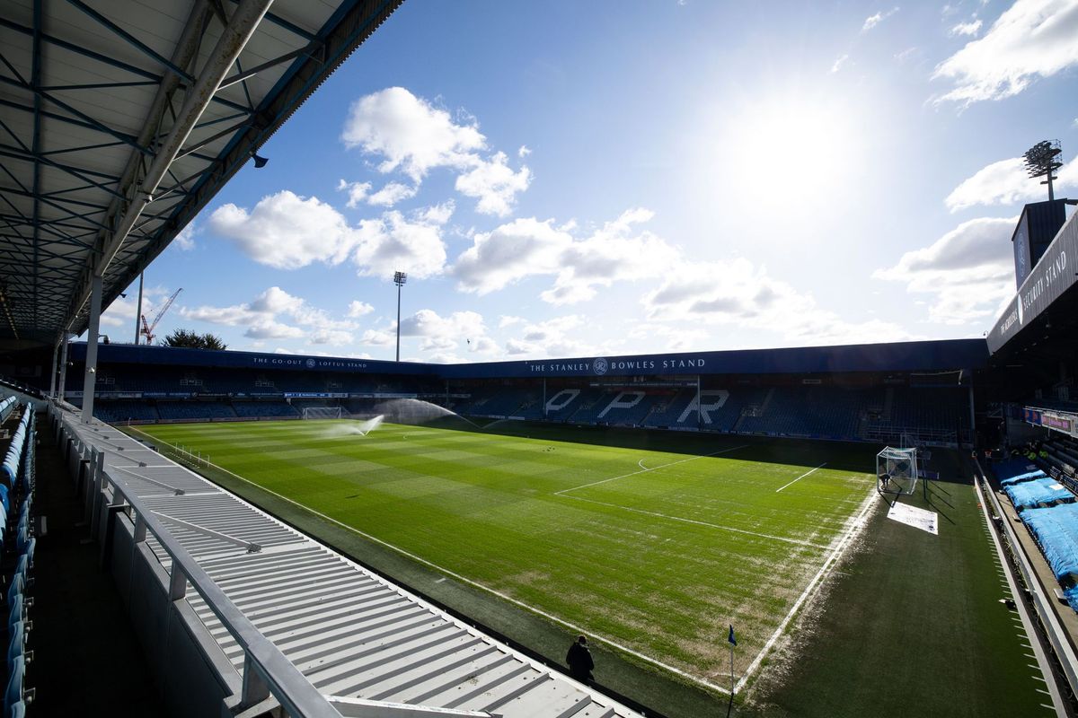 Sunlit empty football stadium with green pitch and blue seating under a bright sky