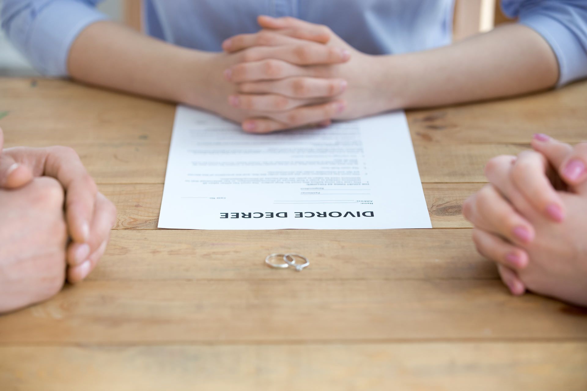 Divorce decree on a wooden table with wedding rings. Hands clasped on both sides, with document in front.