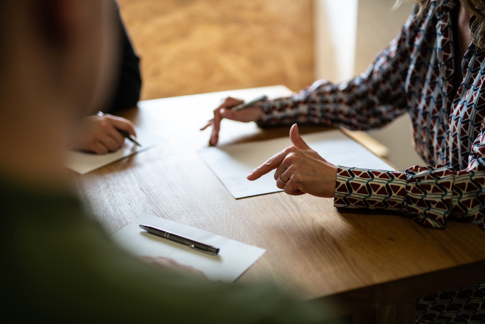 People at a wooden table, one pointing at a paper, another writing. Sunlight warms the scene.