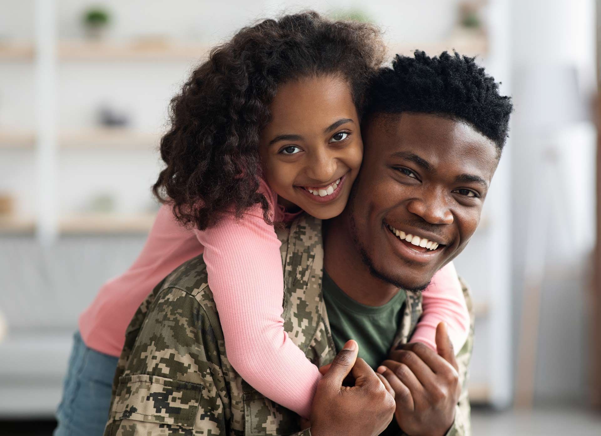 Smiling person in camouflage uniform is embraced by a smiling girl; indoor setting.
