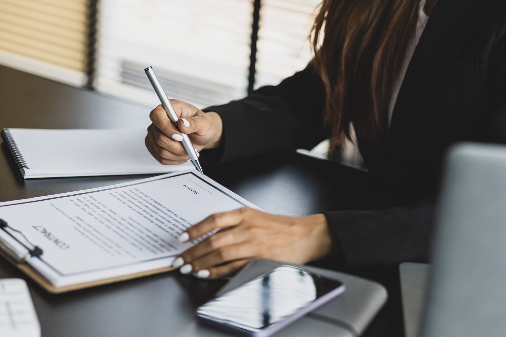 Person signing a contract at a desk, with a pen, phone, and laptop visible.
