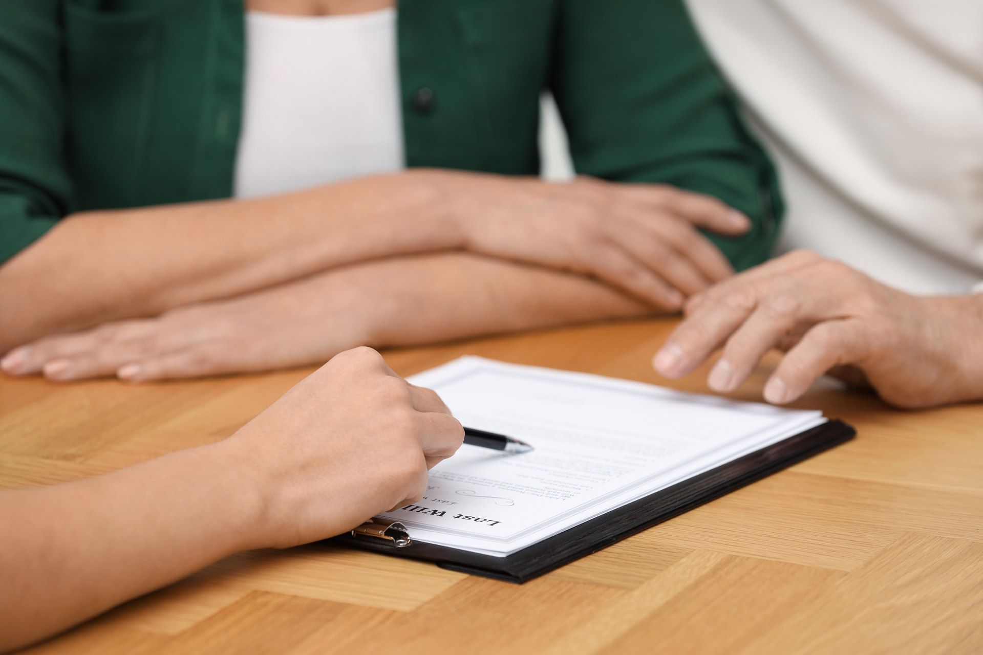Person signing a document on a clipboard at a table, with two other people present.