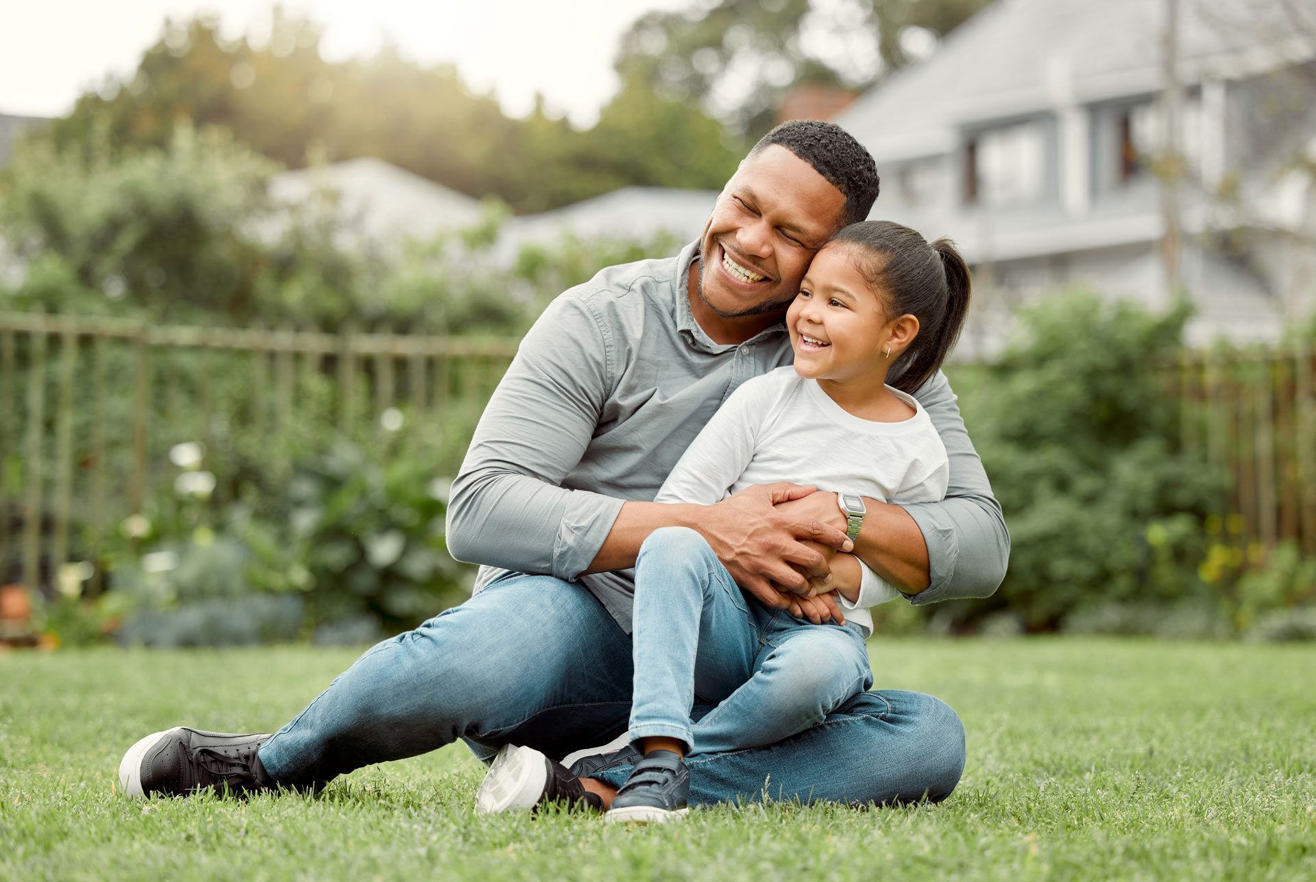 Man and girl sitting on grass, smiling. Man has arms around girl, outdoors.