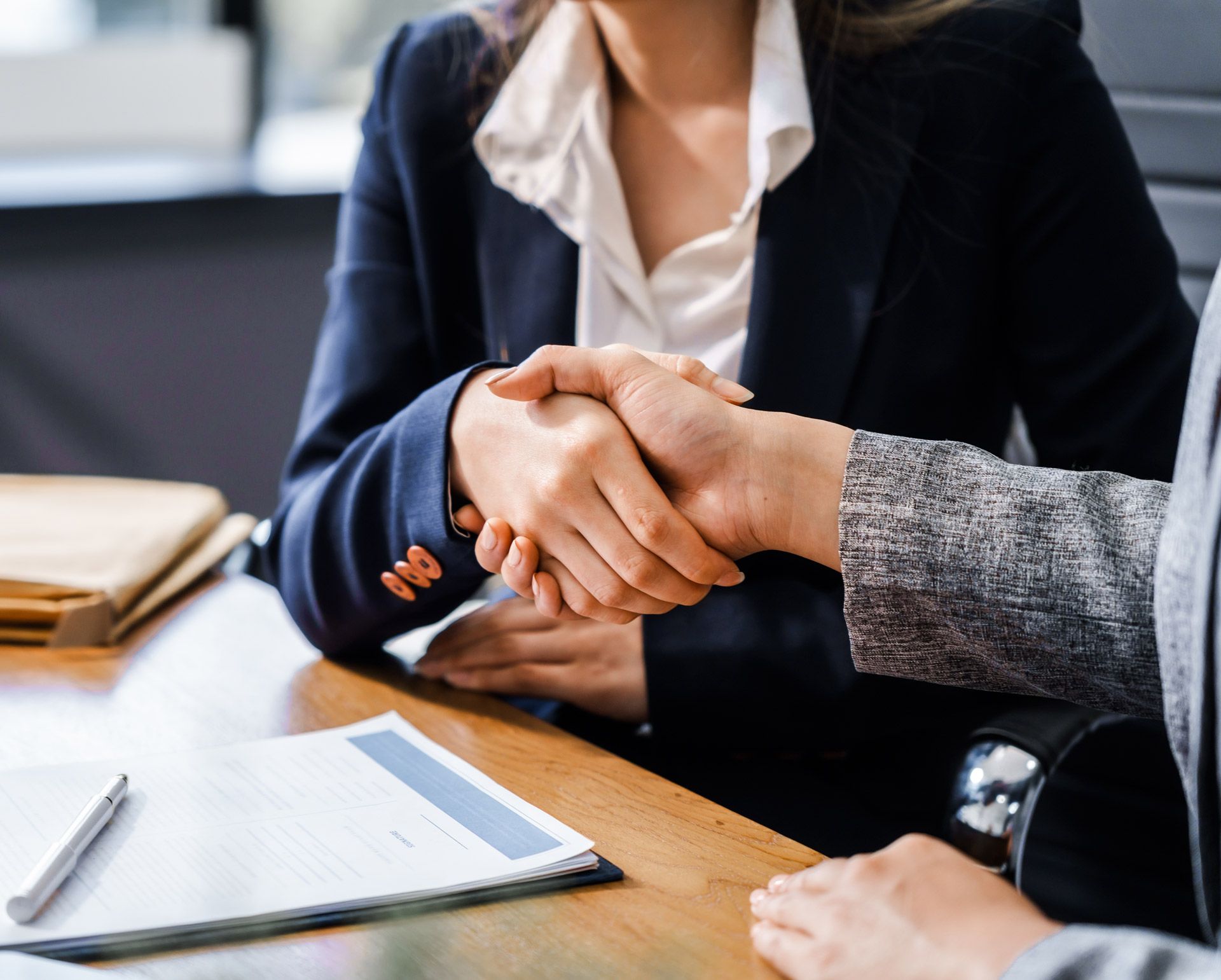 Two people in business suits shaking hands over a desk.