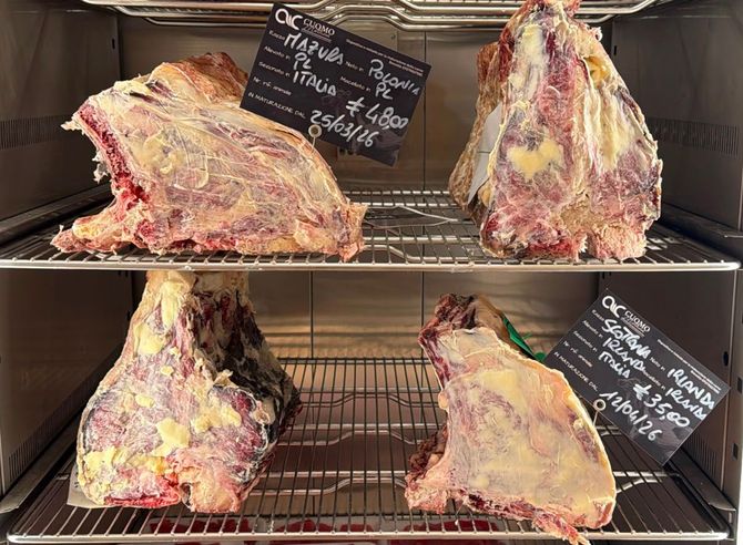 Sezioni di carne cruda tagliate esposte su griglie metalliche in una vetrina da macelleria, con cartellini dei prezzi.