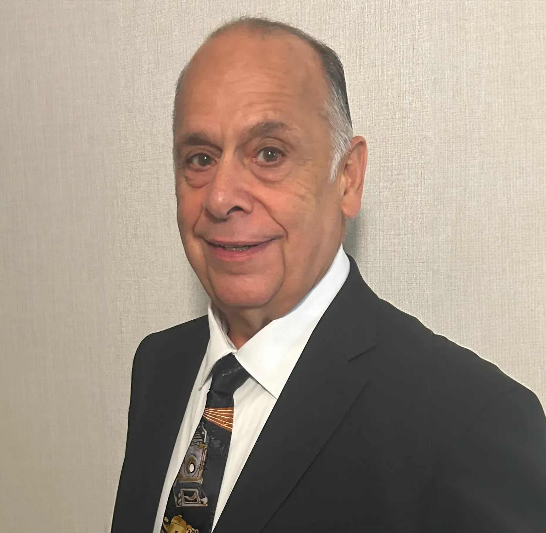 Man in a black suit and patterned tie, smiling against a plain beige background
