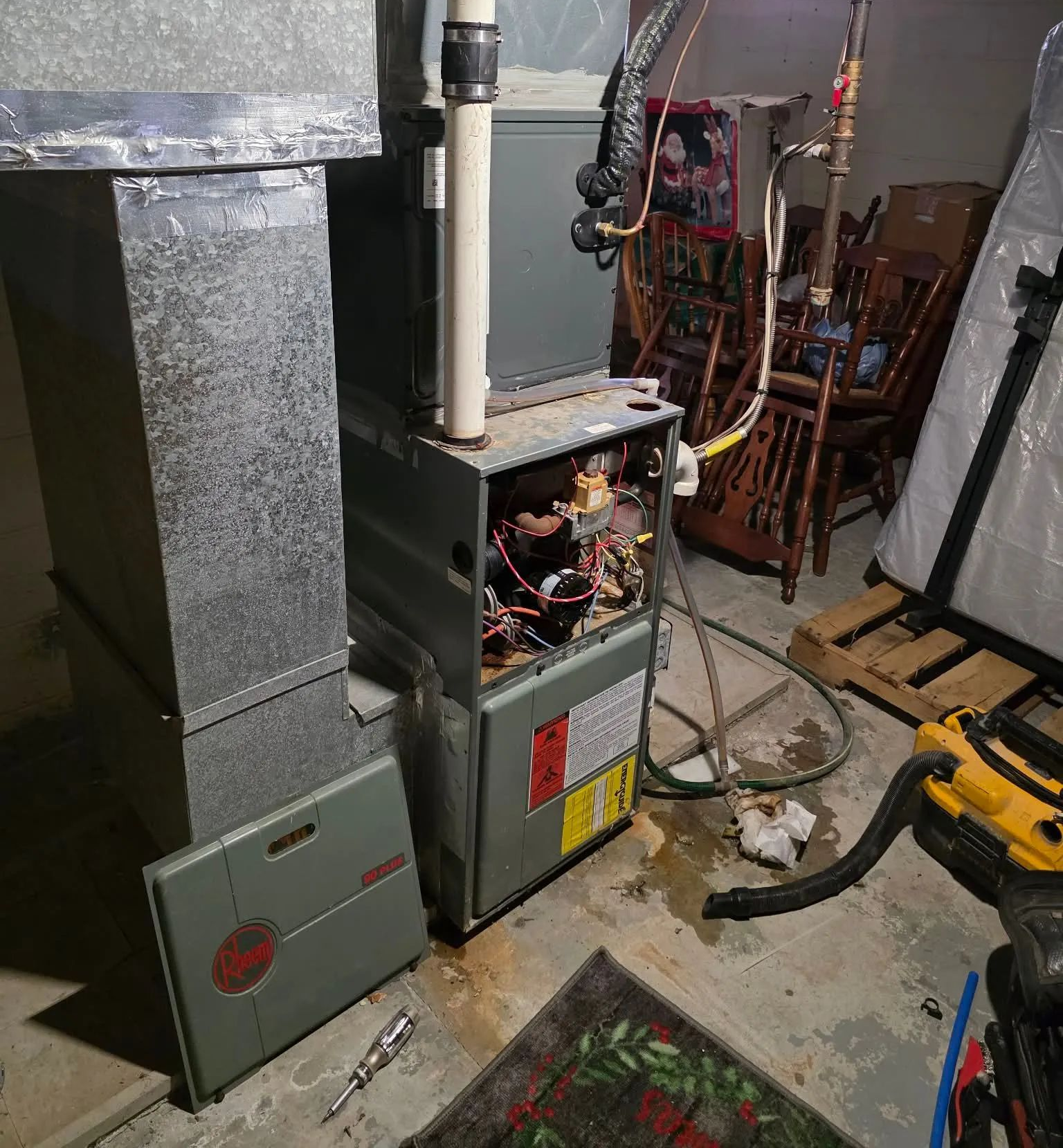 An open furnace with its metal front panel removed, sitting in a basement near tools and stacked furniture.