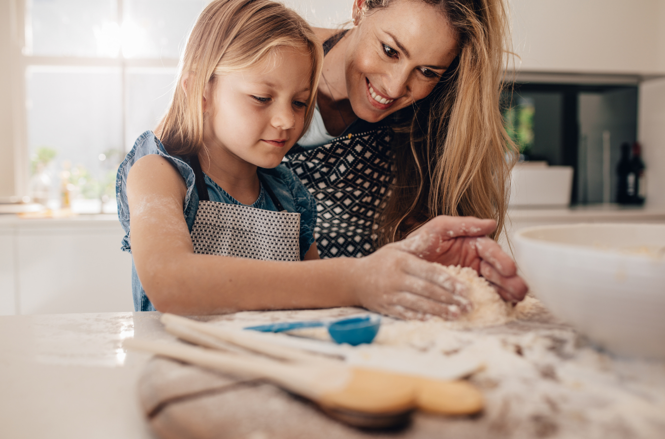 A young girl and a woman with long hair smiling while kneading dough together on a kitchen counter.