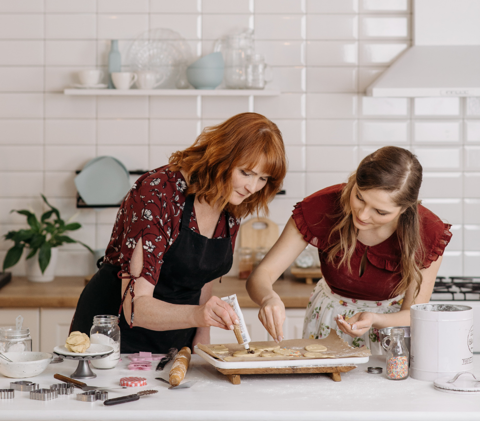 Two women decorate cookies in a kitchen, one piping icing, the other adding sprinkles.