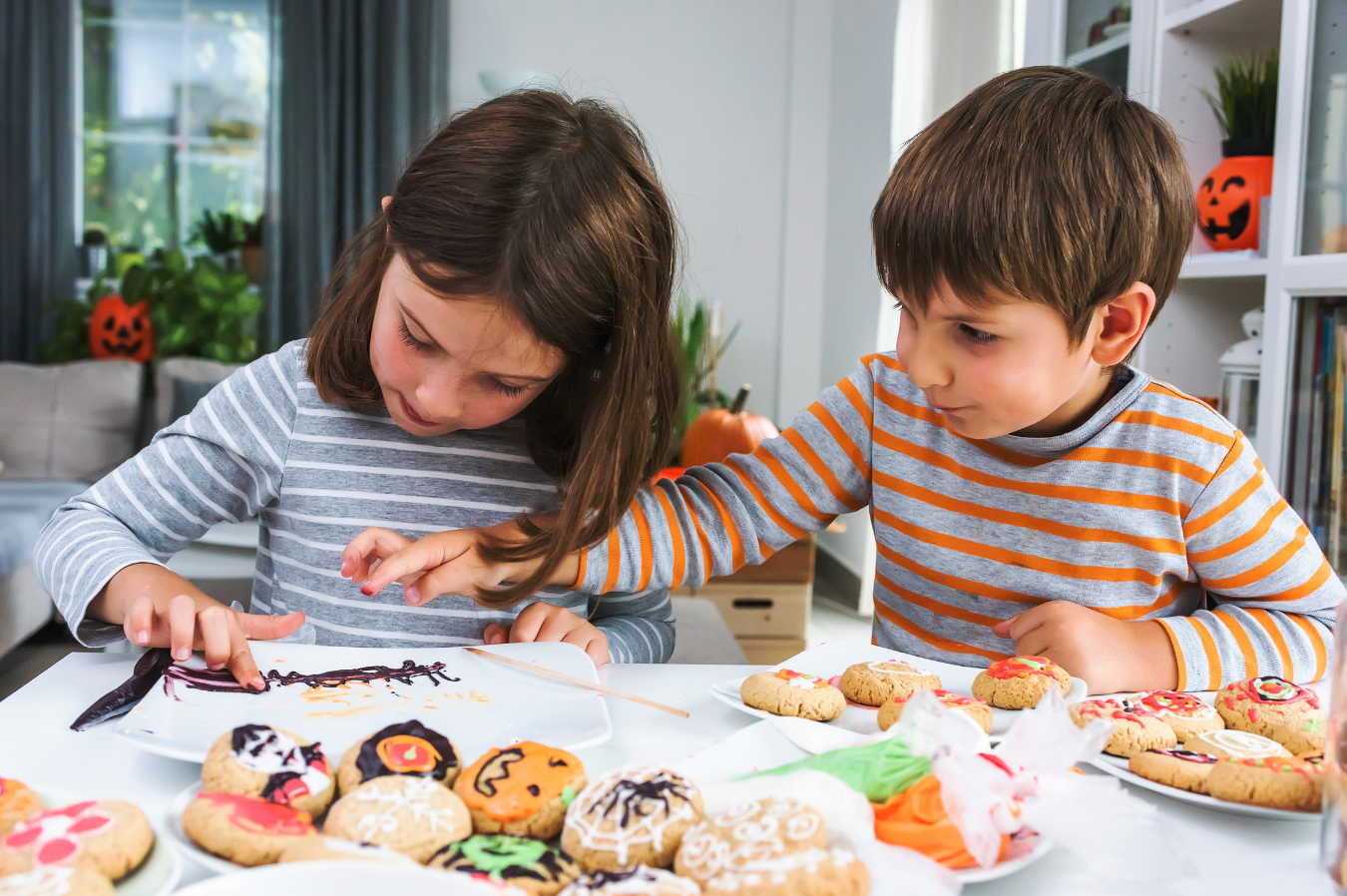 Two children decorate Halloween-themed cookies with icing and sprinkles at a table.