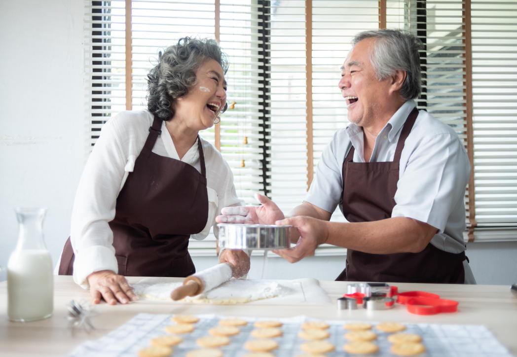 Two people wearing brown aprons laugh while baking together in a bright kitchen with dough and flour on the counter.