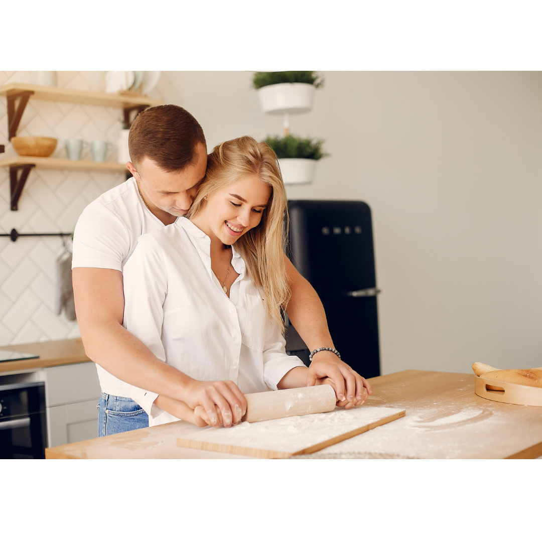 A couple in white shirts smiles while using a rolling pin on dough on a wooden kitchen countertop.
