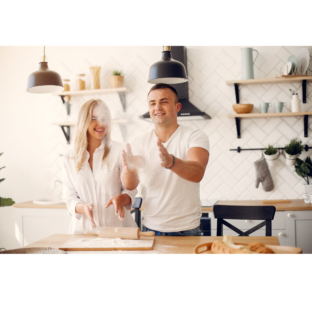 A couple in white shirts laughing while clapping hands covered in flour in a bright kitchen.