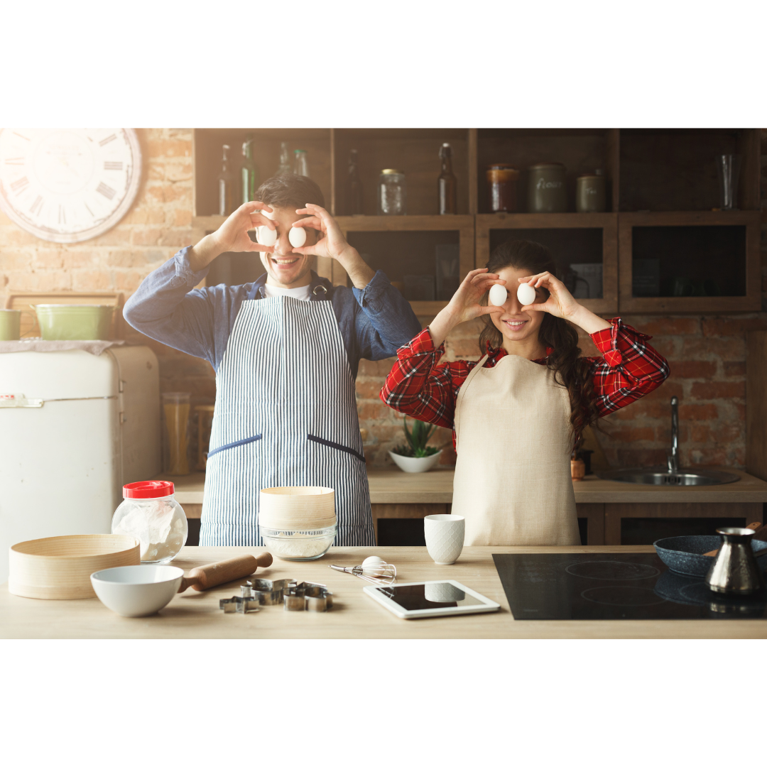 A smiling couple in aprons holds eggs in front of their eyes in a rustic kitchen while preparing to bake.