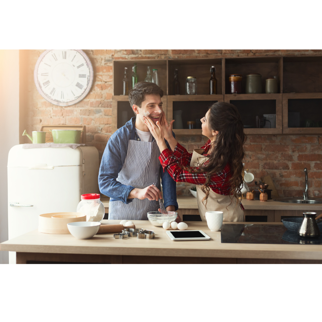 A couple in aprons laughing together while baking in a kitchen, with one person playfully touching the other's face.