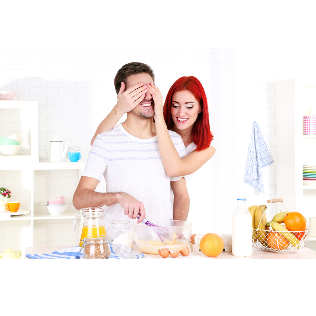 A woman with red hair playfully covers a man's eyes in a bright kitchen while he prepares food at a counter.