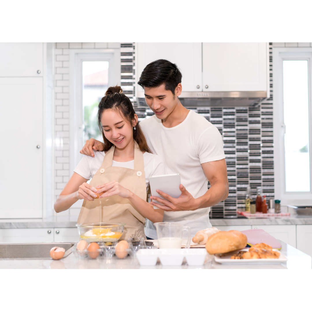 A couple in aprons stand in a modern kitchen, smiling as they look at a tablet while preparing food with eggs and bread.