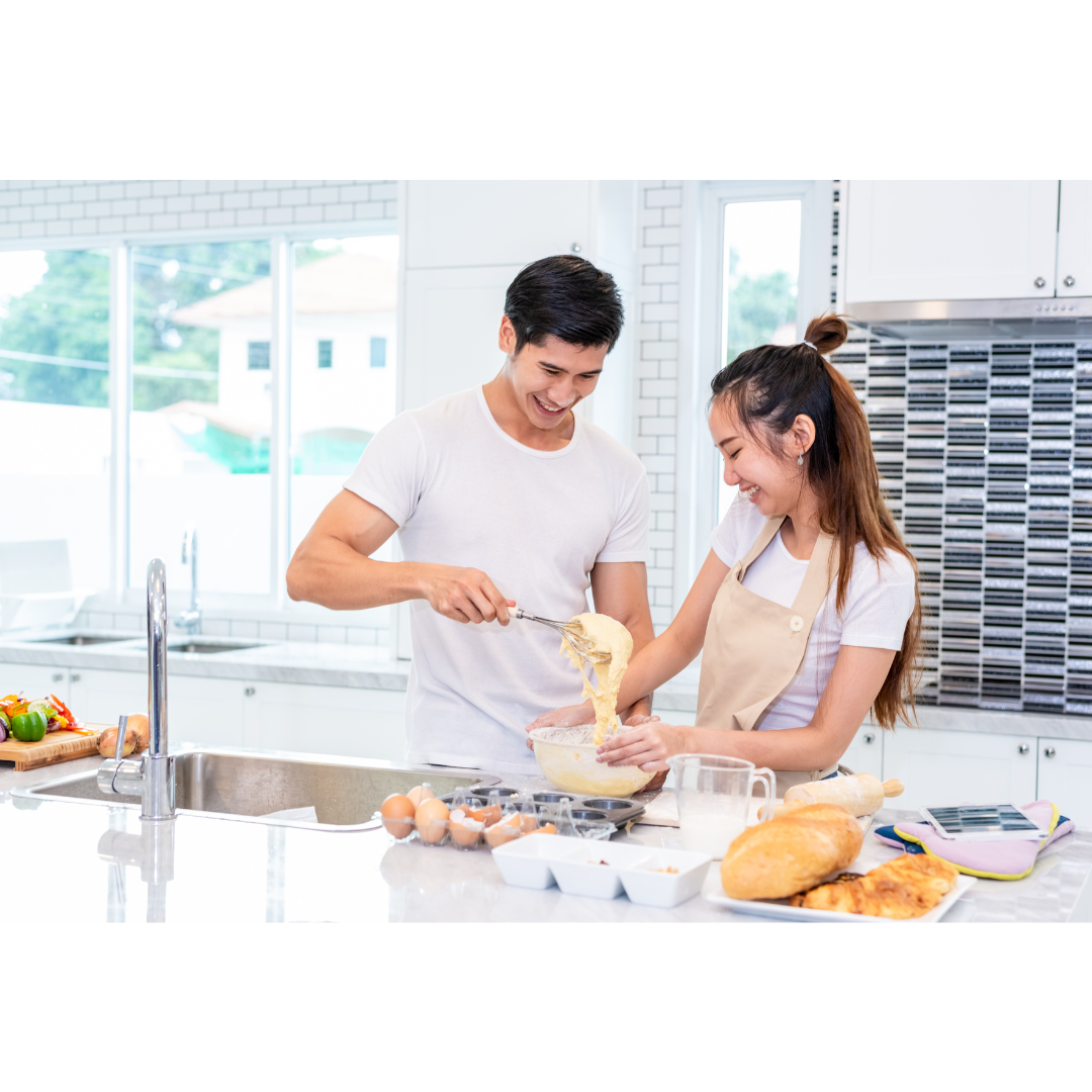 A smiling couple in a white-tiled kitchen, mixing batter together in a bowl.