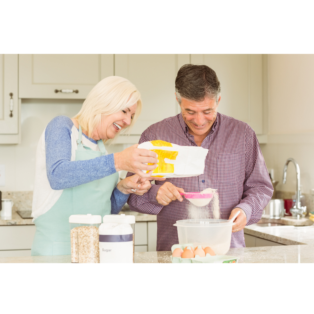 Two people in a kitchen laughing while sifting flour into a bowl together.