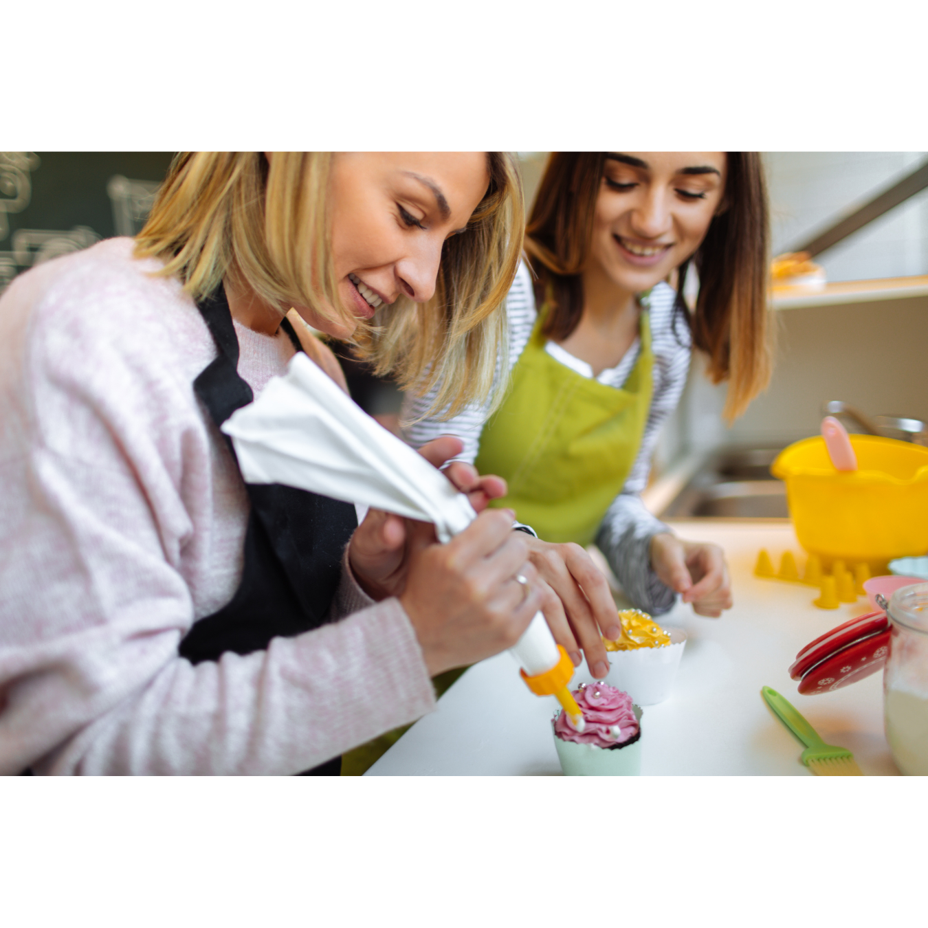 Two people wearing aprons decorate a cupcake with frosting using a piping bag in a kitchen setting.