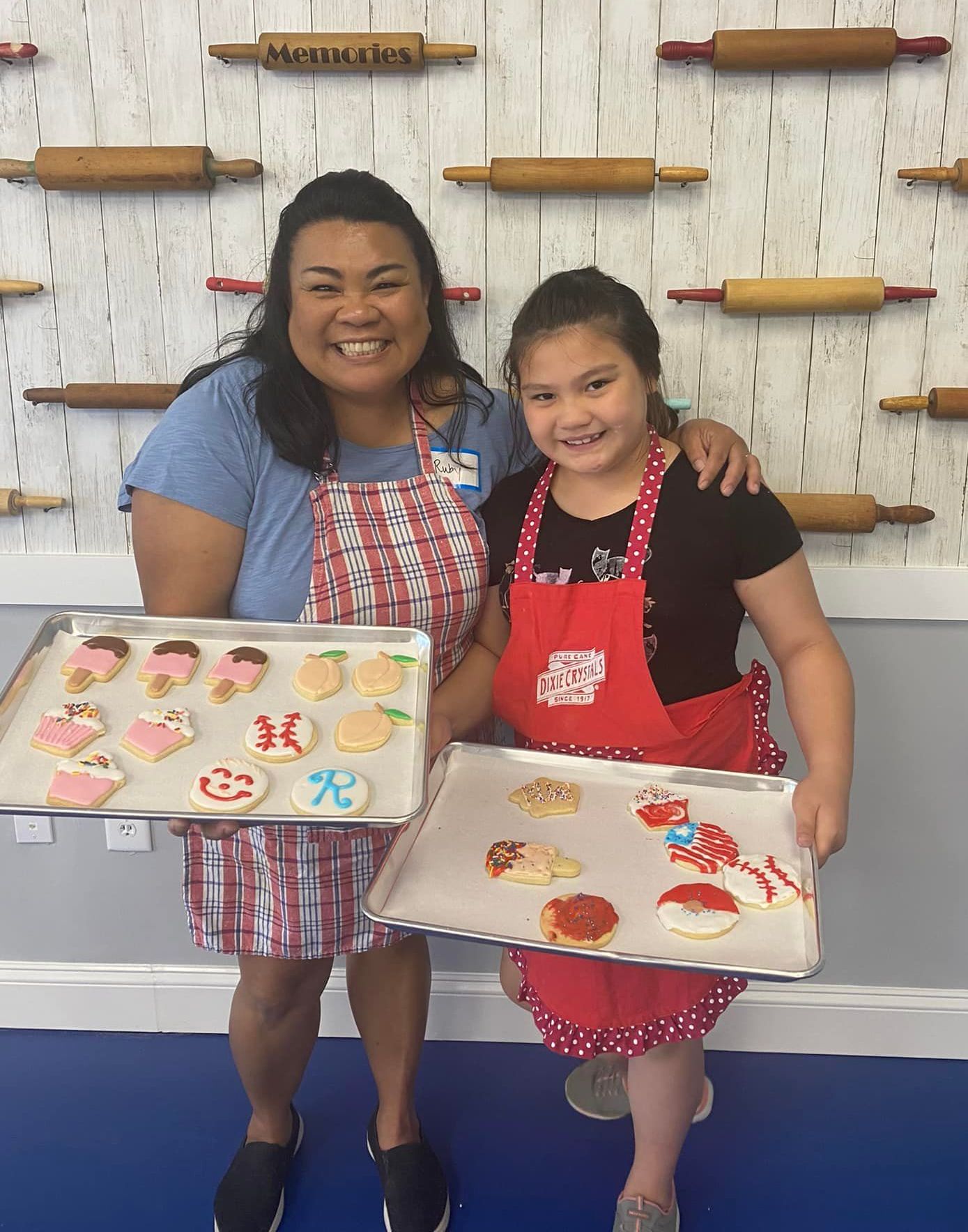 Two people wearing aprons stand in front of a wall decorated with rolling pins, holding trays of decorated cookies.