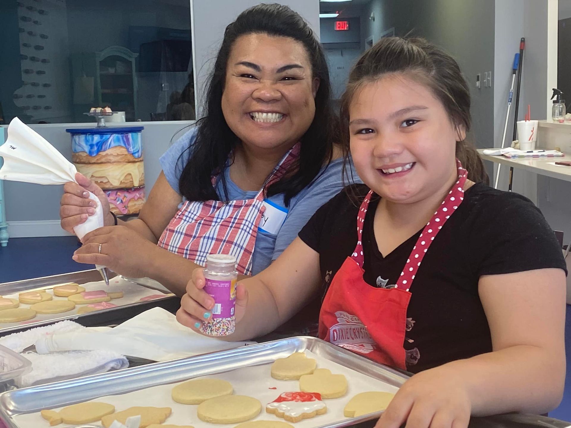 Two people wearing aprons smile while decorating cookies on trays in a kitchen setting.