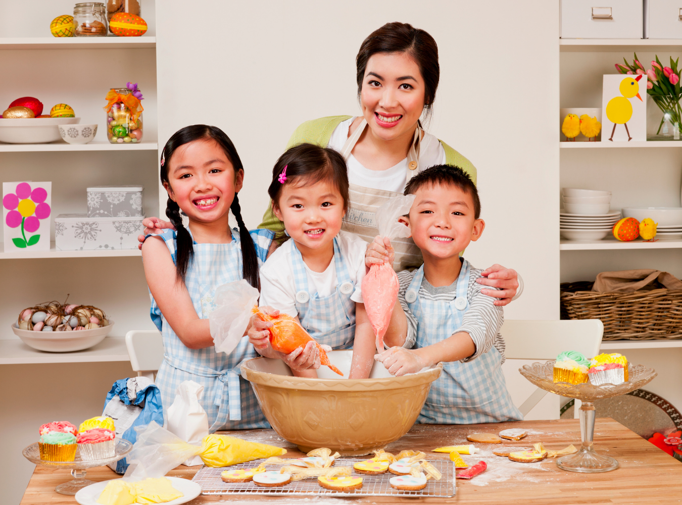 A smiling adult and three children in matching aprons decorate cookies in a bright kitchen.