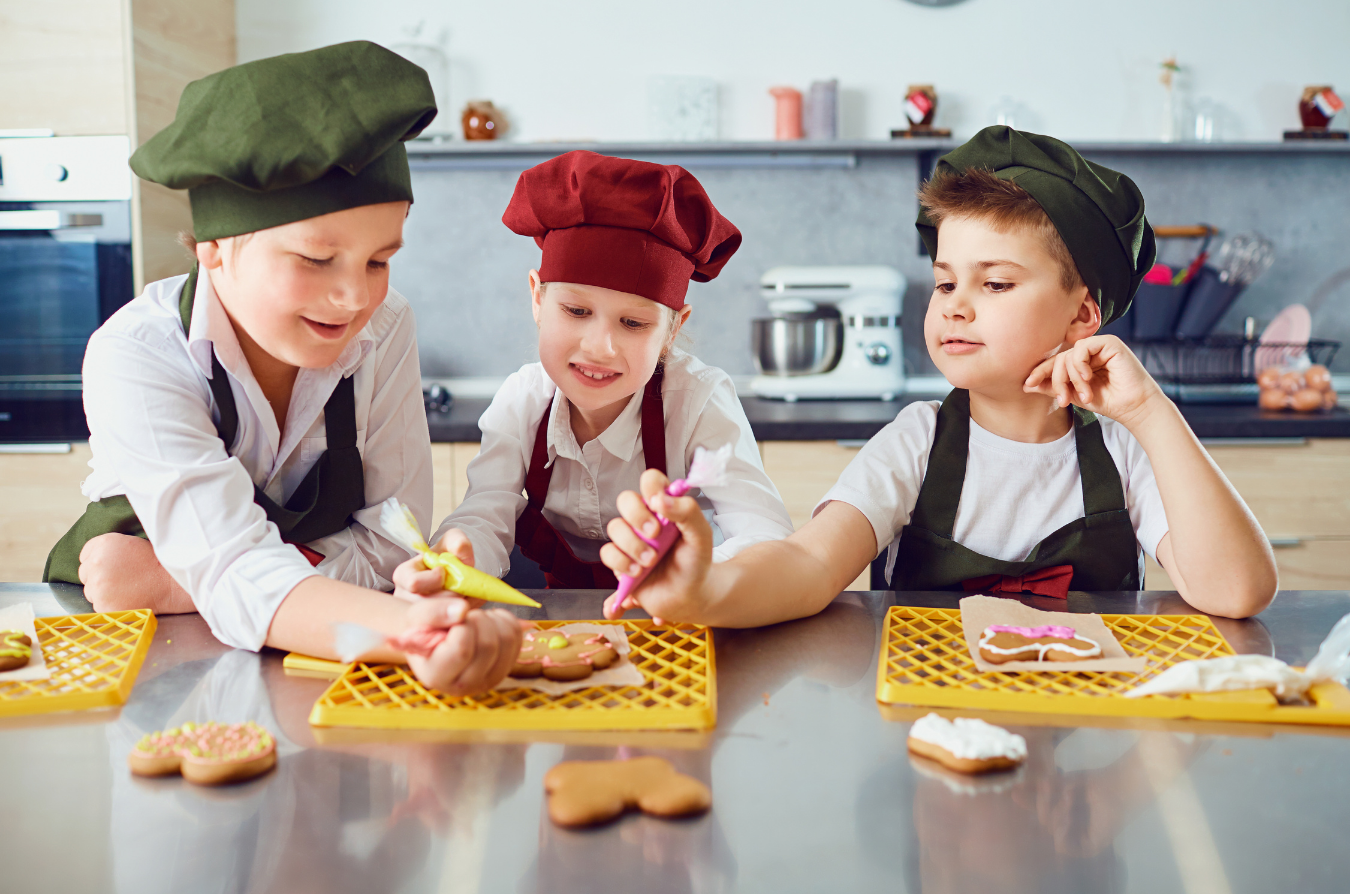 Three children wearing chef hats and aprons decorate gingerbread cookies in a kitchen.