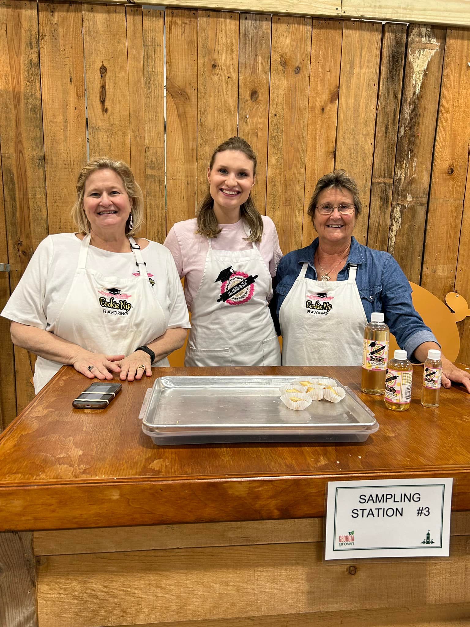 Three people wearing aprons stand behind a wooden counter with a serving tray and bottles at a food sampling station.