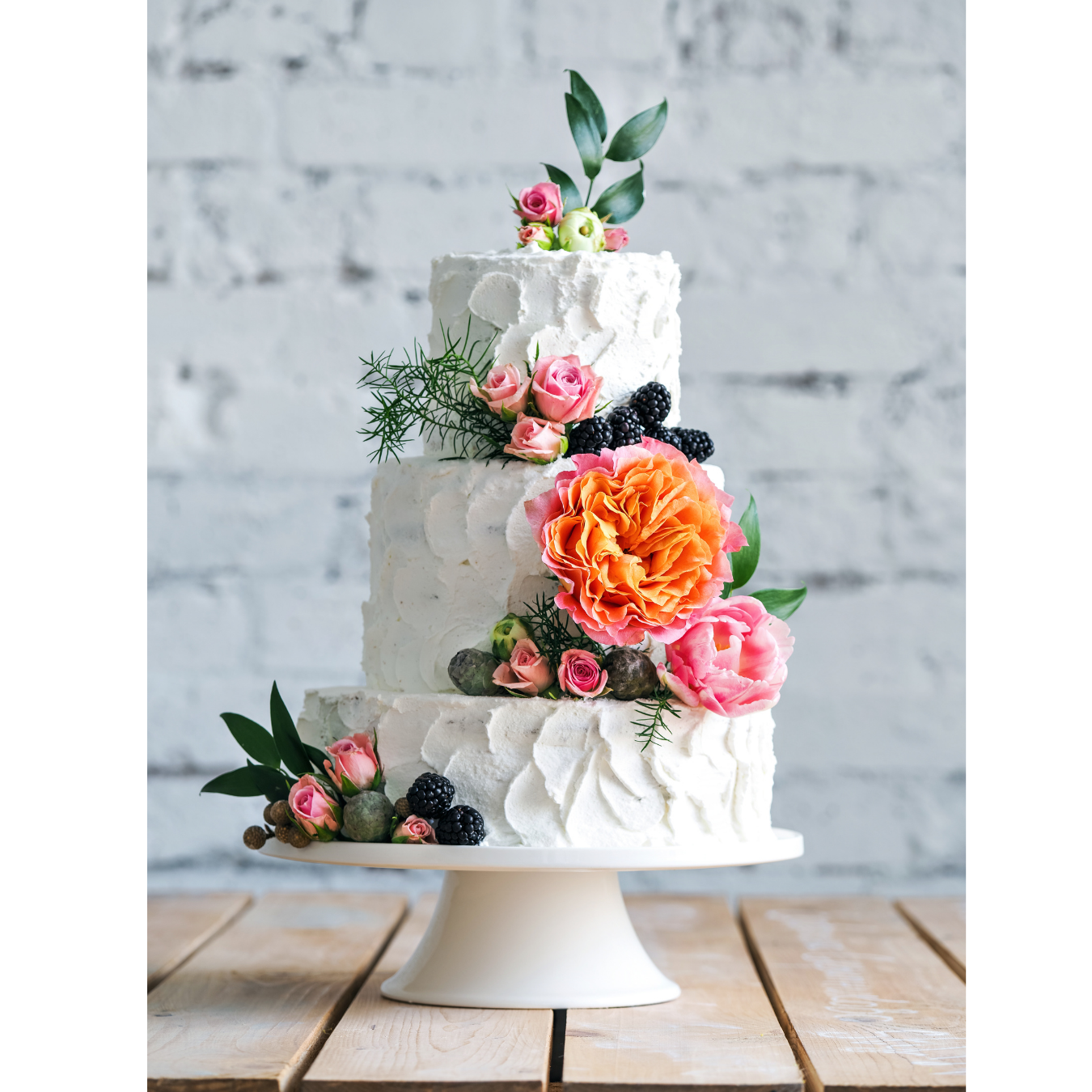 A three-tiered white cake decorated with vibrant orange and pink flowers and fresh blackberries on a white pedestal.