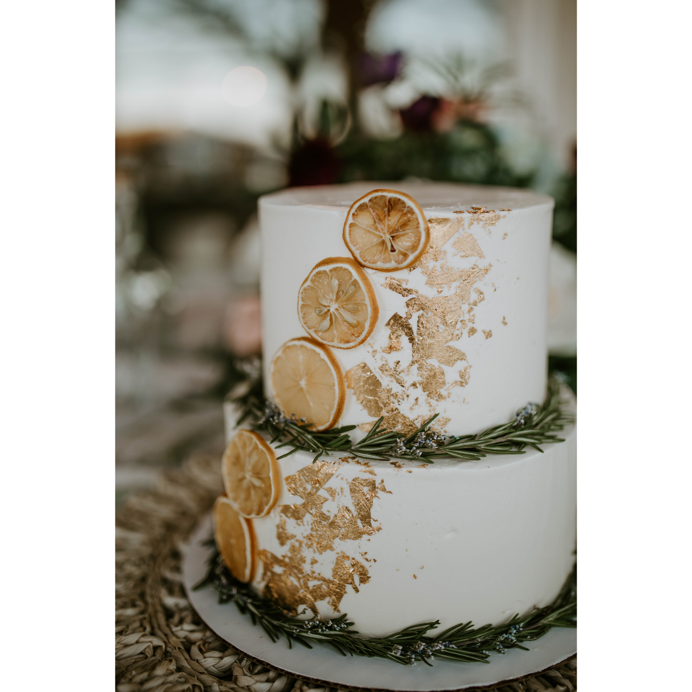 Two-tiered white wedding cake decorated with dried lemon slices, gold leaf accents, and sprigs of fresh rosemary.