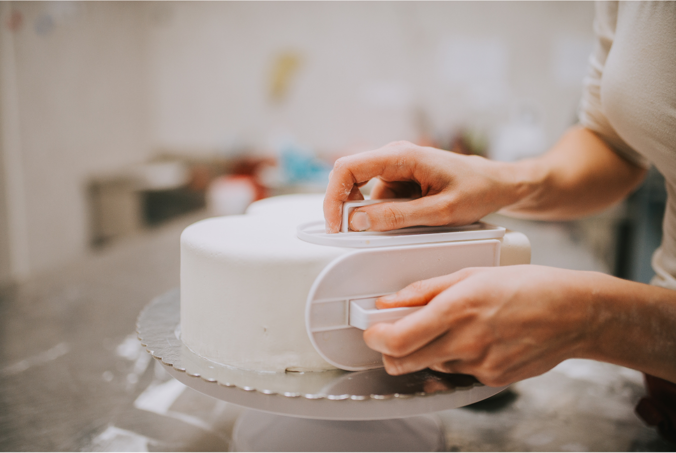 A person uses a white plastic smoothing tool to flatten the fondant frosting on a round white cake.